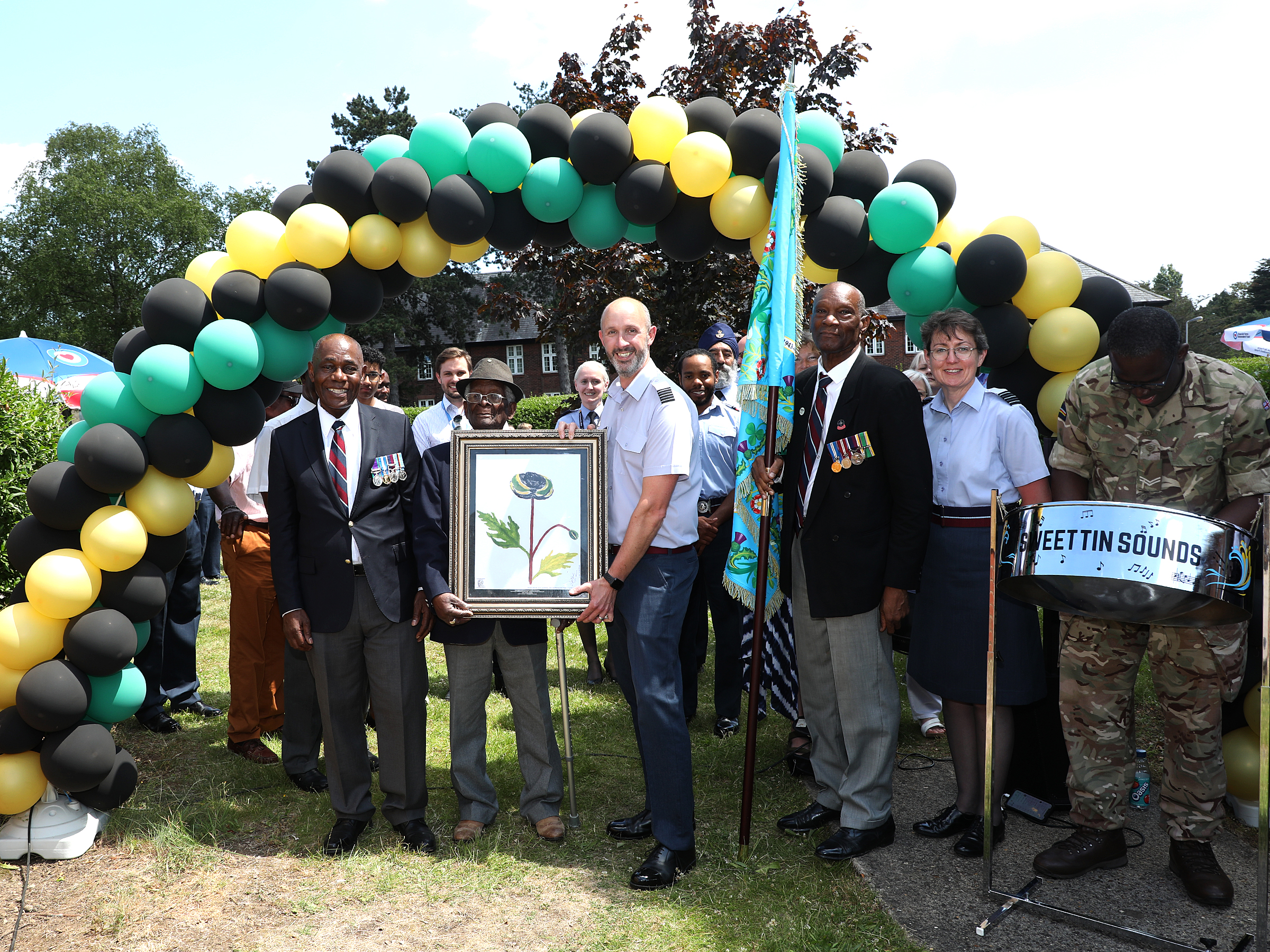 Aviators hold certificate under arc of balloons in the colours of the Jamaican flag, with steel drum player.